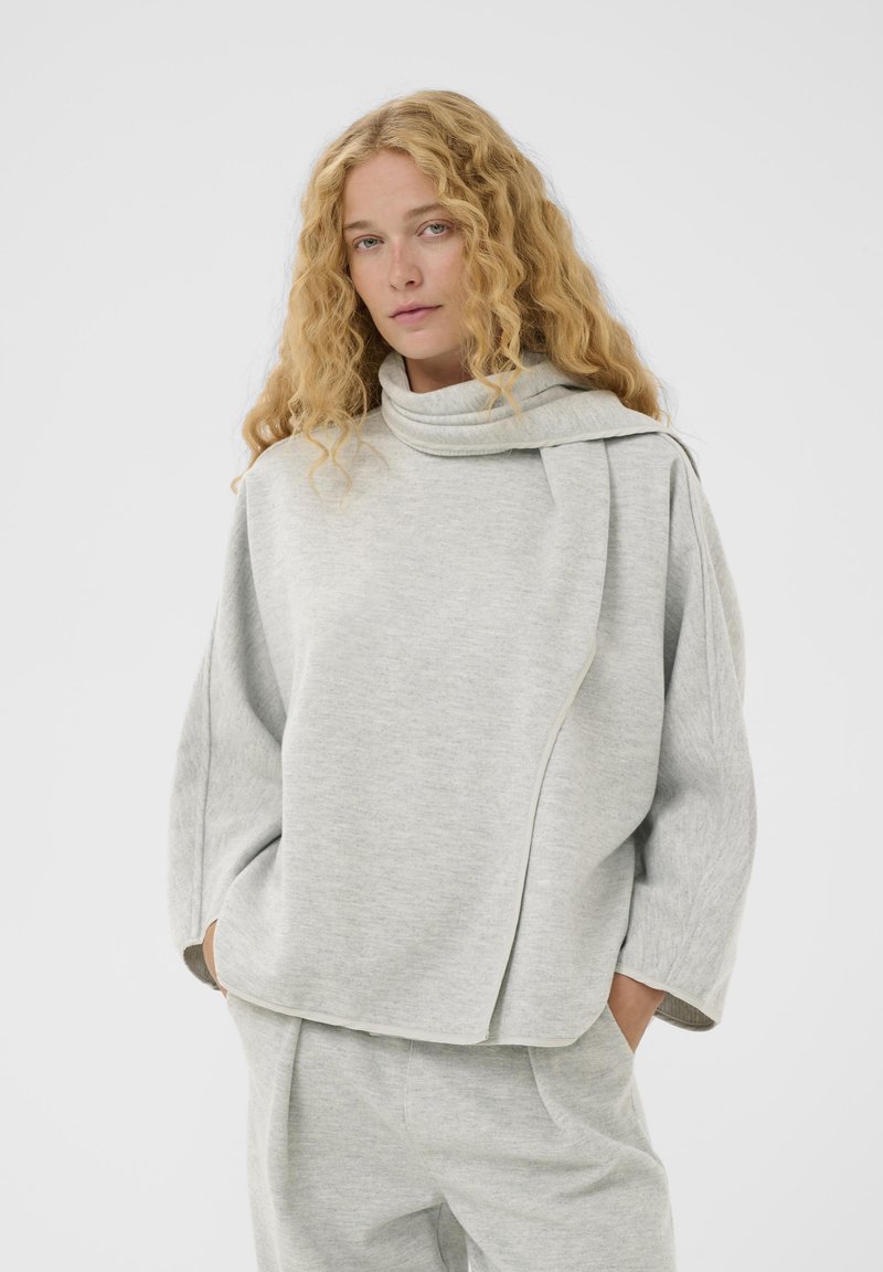 Világosszürke, oversized kapucnis pulóver magas nyakkal, aszimmetrikus dizájnnal és puha tapintással. Széles ujjú és laza szabású.