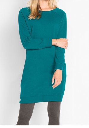 Femme portant une robe-tunique à manches longues bleu sarcelle avec poignets et ourlet côtelés, associée à des leggings gris foncé sur un fond blanc uni.