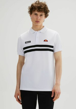 Młody mężczyzna w białej koszulce polo z krótkim rękawem, czarnymi podwójnymi paskami i logo ellesse, w czarnych spodniach, stojący na jednolitym tle.