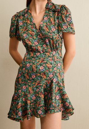 Grøn kort blomsterkjole med orange, pink og blå blomster, puffede korte ærmer og en flæsekant, båret af en stående person.