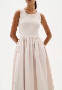 Vestito beige senza maniche con corpino aderente a coste e gonna ampia arricciata indossato da una persona con le mani dietro la schiena.