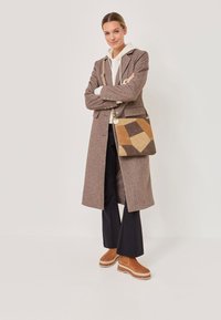 Žena v hnědém dlouhém kabátu, bílém mikině s kapucí, černých kalhotách a béžových botách, držící křížovou tašku v patchwork designu béžové a hnědé barvy, stojí se zkříženýma rukama.