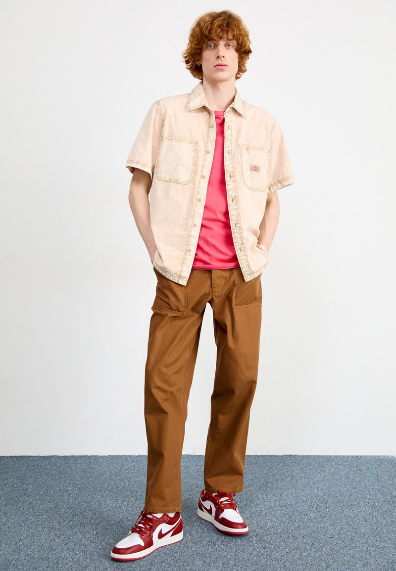 Chemise beige à manches courtes avec deux poches poitrine, t-shirt rose en dessous, pantalon marron plissé et basket rouge et blanche.