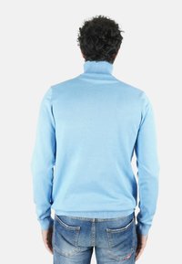 Maglione a collo alto color azzurro chiaro a coste con maniche lunghe, abbinato a jeans blu sbiaditi, visto da dietro. Tessuto liscio, vestibilità casual.