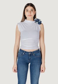 Top bianco senza maniche con collarino alto e texture arricciata, caratterizzato da un accento decorativo di un fiore blu sulla spalla. Indossato con jeans blu.
