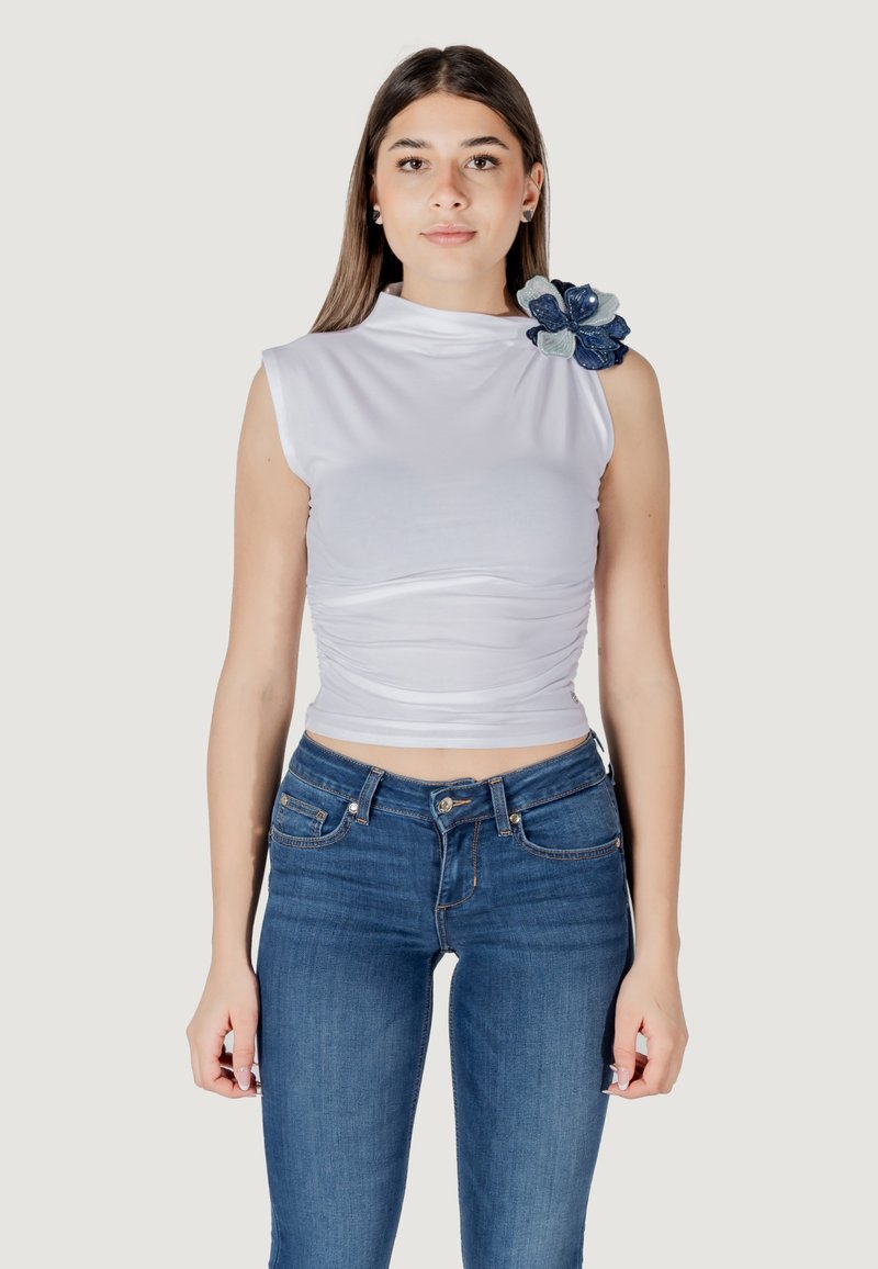 Top bianco senza maniche con collarino alto e texture arricciata, caratterizzato da un accento decorativo di un fiore blu sulla spalla. Indossato con jeans blu.