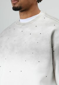 Rondehals sweater in lichtgrijs met een verloop effect. Voorzien van verspreide blauwe en zwarte decoratieve studs, gemaakt van zachte stof met een gladde textuur.