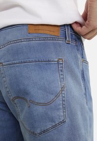 Light blue denim jeans featuring a textured surface, a brown leather label at the waistband, and contrasting orange stitching on the pockets.