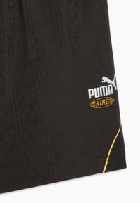 Schwarzer Stoff mit dezenten, erhabenen geometrischen Mustern, weißes Puma-Logo, gelber Schriftzug "King" innerhalb eines Ovals und ein gebogener gelber Streifen nahe dem Saum.