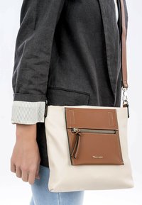 Hnědá crossbody taška s krémovým tělem, hnědou koženou kapsou, stříbrným zipem a kontrastním prošíváním; hladký povrch a nastavitelný popruh.