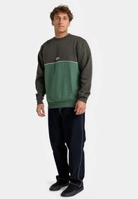 Sweatshirt verde e cinza escuro com gola redonda, faixa horizontal contrastante e pequeno logotipo. Combinado com jeans escuros e sapatos pretos.