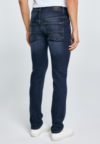 Muškarac u tamnoplavim trapericama uskog kroja i bijelim tenisicama stoji okrenut leđima prema kameri, na jednostavnoj svijetloj pozadini.