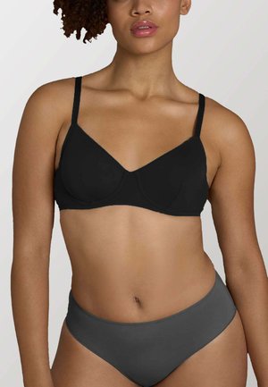 Donna che indossa un reggiseno nero e biancheria intima grigia, in piedi contro uno sfondo chiaro e uniforme, mostrando il busto superiore e i fianchi.