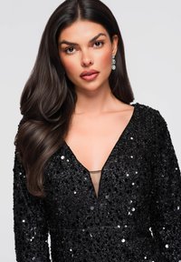 Robe noire à sequins avec manches longues et décolleté plongeant en V. Comprend un panneau transparent au niveau de la poitrine et un travail de perles détaillé pour la texture.