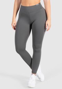 Mujer con leggings de talle alto en gris oscuro y zapatillas blancas, de pie con las piernas cruzadas contra un fondo neutro.