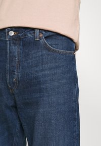 Jeans in denim blu scuro con vestibilità classica, caratterizzati da cuciture visibili, chiusura con bottone e tasche anteriori. Tessuto morbido e materiale resistente.