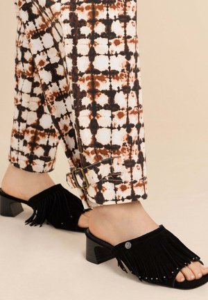 Jambes portant un pantalon à motifs avec un détail de boucle roulé au-dessus de sandales à talons noires avec franges et orteils ouverts sur un sol beige.