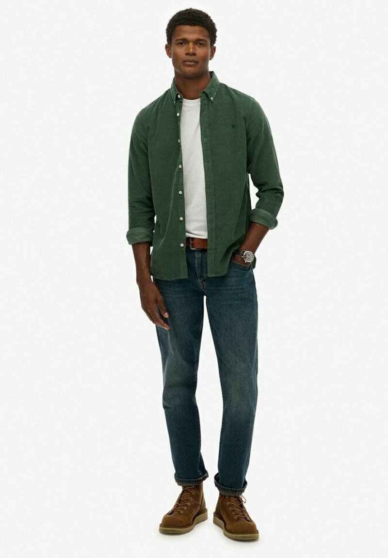 Groene button-up shirt, witte t-shirt eronder, blauwe jeans, bruine veterschoenen en een horloge. Glad materiaal en casual pasvorm.