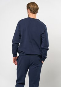 Námornícky modrý sveter s dlhými rukávmi, okrúhlym výstrihom a rebrovanými manžetami. Materiál sa zdá byť mäkký, s hladkou textúrou a minimalistickým dizajnom.