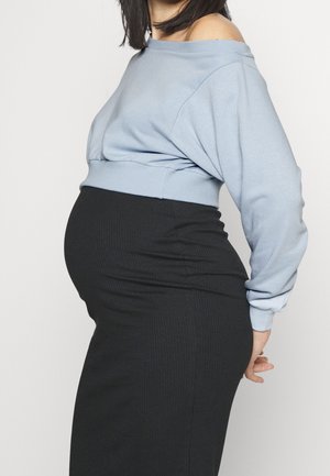 Gravid person iført en lyseblå off-shoulder sweater og en tætsiddende sort nederdel, der poserer sidelæns mod en ensfarvet baggrund.