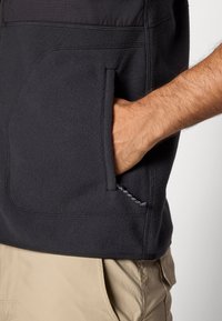 Crna fleece jakna s teksturiranom površinom, s bočnom džepom i vidljivim detaljima šivanja. Nosi se preko bež hlača.