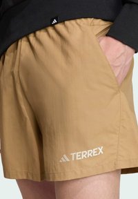 Bež Terrex kratke hlače od lagane tkanine, s bočnom džepom i bijelim logotipom blizu rubova. Elastičan pojas.