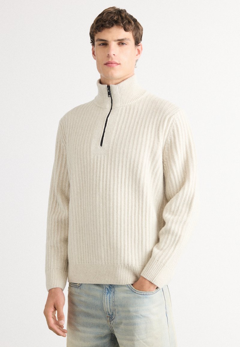 Helle beige Strickjacke mit hohem Kragen und halbem Reißverschluss. Verfügt über ein strukturiertes Rippdesign und lange Ärmel. Getragen mit hellblauen Jeans.