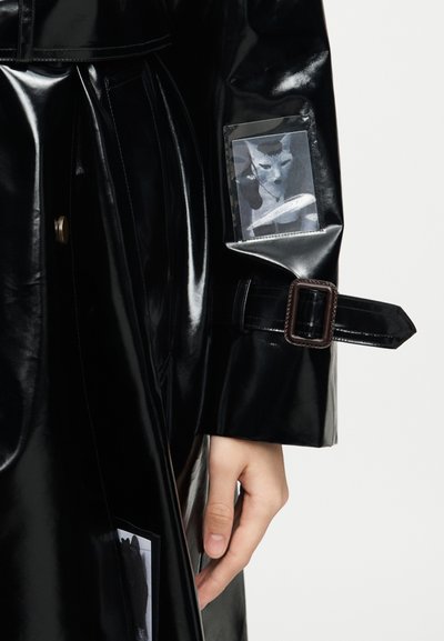 Γυαλιστερό μαύρο μανίκι παλτού-τραντσκότ με ρυθμιζόμενο ιμάντα και προσαρτημένη μικρή φωτογραφία γάτας στο αντιβράχιο, με το χέρι ορατό κάτω από το μανίκι.