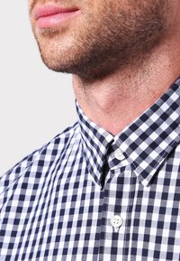 Ingrandimento della parte inferiore del viso e del collo di un uomo con leggera barba di tre giorni, che indossa una camicia a quadri bianca e nera.