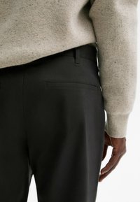 Pantalon noir en tissu lisse avec une simple poche arrière. Porté avec un pull gris clair avec de subtils flocons.