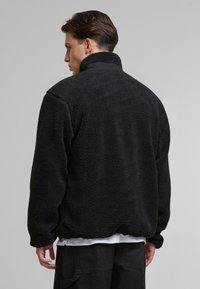Svart fleece-jakke med høy krage, strukturert stoff og en avslappet passform. Har lange ermer og et uformelt design som er egnet for lagdeling.