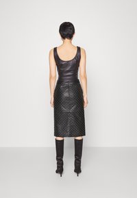 Femme aux cheveux courts portant un haut noir en cuir sans manches, une jupe en cuir noire cloutée et des bottes noires à talons, debout face à un mur uni.