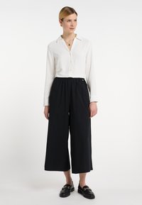 Witte blouse met lange mouwen en knoopsluiting, gecombineerd met zwarte wijde broek. De blouse heeft een kraag; de broek heeft een elastische tailleband.