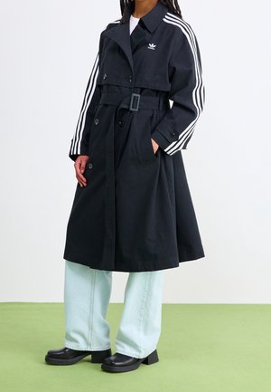 Personne portant un trench-coat noir Adidas ceinturé avec des bandes blanches sur les manches, un jean large bleu clair et des chaussures noires à talons, debout sur un sol vert.