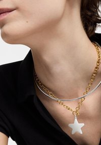 Collana in metallo con una catena dorata e un ciondolo a forma di stella liscia in argento, indossata su una camicia scura con colletto.