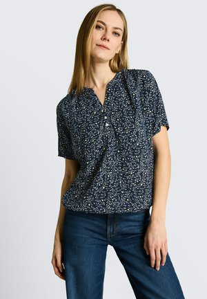 Vrouw draagt een donkerblauwe korte mouwen blouse met witte en gele bloemenprint, gecombineerd met blauwe jeans, staand tegen een effen achtergrond.