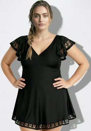 Frau mit geflochtenem Haar, die ein schwarzes V-Ausschnitt-Kleid mit Spitzenärmeln und Saum trägt, steht mit den Händen in die Hüften gestützt vor einfachem Hintergrund.