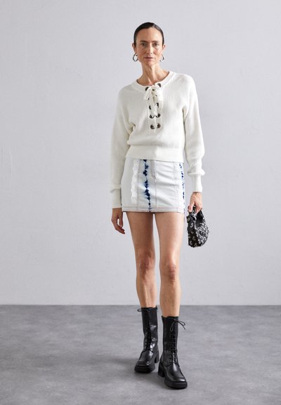 Pull blanc côtelé avec des détails lacés, associé à une jupe mini en denim avec des motifs tie-dye bleus. Bottines noires à lacets et un petit sac.