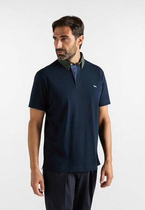 Uomo con barba che indossa una polo blu navy a maniche corte con colletto a strati verde e blu, in piedi davanti a uno sfondo bianco uniforme.