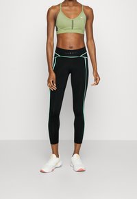 Sportowy top w oliwkowym kolorze z centralnym wycięciem, w połączeniu z czarnymi legginsami z zielonymi akcentami. Buty są białe z pomarańczowymi detalami.