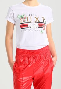 Camiseta de algodón blanca con mangas enrolladas, que presenta gráficos florales, texto dorado y detalles de rayas rojas, combinada con pantalones jogger rojos brillantes.