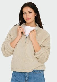 Pull en polaire beige avec un col zippé mi-hauteur et des épaules tombantes. Présente un tissu texturé et des poignets côtelés. Porté par-dessus une chemise blanche.