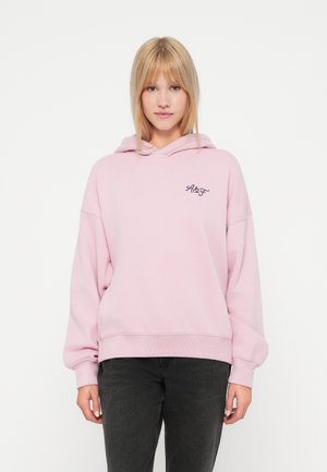 Šviesiai rožinė didelio dydžio džemperis pagamintas iš minkštos medžiagos, turintis raištelius ant gobtuvo, lanksčias rankogalius ir mažą siuvinėtą logotipą ant krūtinės.