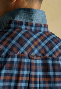 Karo krekls ar zilu un brūnu pārbaudi, džinsu apkakli, auduma birku ar "JOULES" un teksturētu apdari. Attēlots aizmugurējais skats.