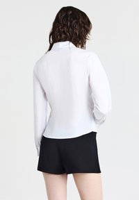Una donna con i capelli bagnati indossa una blusa bianca attillata a maniche lunghe e pantaloni corti neri, vista da dietro su uno sfondo neutro.