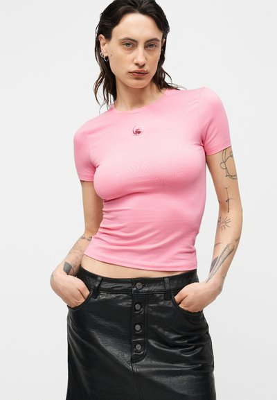 Νεαρό άτομο με σκούρα μαλλιά μέχρι τους ώμους, φοράει εφαρμοστό ροζ μπλουζάκι και μαύρη δερμάτινη φούστα με κουμπιά, τα χέρια στις τσέπες, τατουάζ εμφανή.