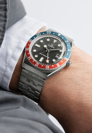 La montre à quartz Timex Q GMT présente un cadran noir, une lunette rouge et bleue, un bracelet en acier inoxydable argenté et des index lumineux.