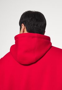 Sudadera roja con un alto cuello y capucha ajustable. El material parece suave y texturizado, con costuras visibles a lo largo de las costuras.