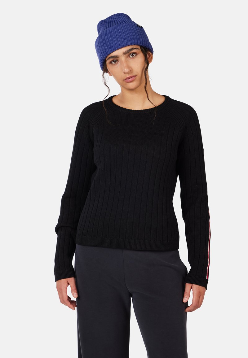 Schwarzer gerippter Strickpullover mit langen Ärmeln, Rundhalsausschnitt und rot-weißen Streifen am linken Ärmel. Kombiniert mit dunklen Hosen. Blauer Strickmütze.