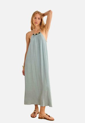 Mujer con vestido maxi azul claro sin mangas con escote decorado con cuentas, combinando sandalias planas con cuentas y pulsera dorada, de pie con el brazo levantado.